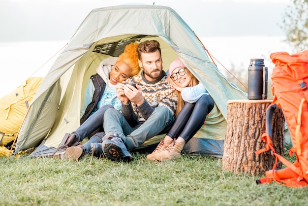 湖の近くの屋外レクリエーション中にテントに座って一緒にウォーミングアップするセーターを着た友人の多民族グループ