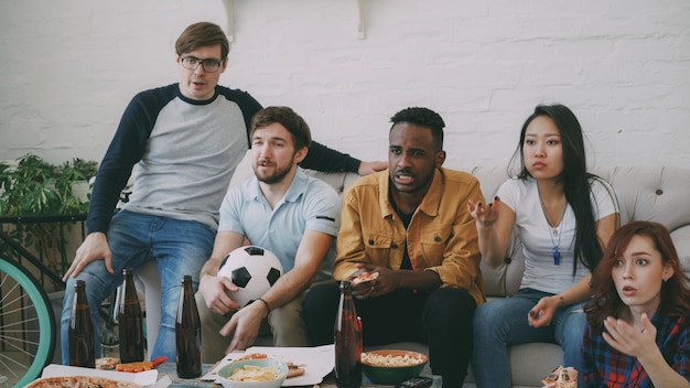 Gruppo multietnico di amici appassionati di sport che guardano il campionato di calcio in tv insieme mangiando pizza e bevendo birra a casa