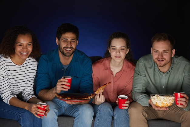 Multi-ethnic group of friends watching movies at home while eating snacks and popcorn sitting on big sofa in dark room