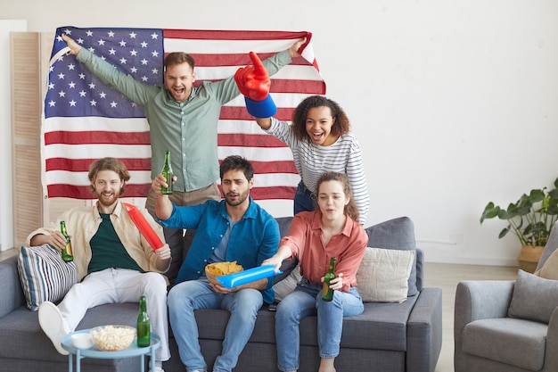 Gruppo multietnico di amici che guardano partite di sport e tifano emotivamente mentre si tiene la bandiera americana