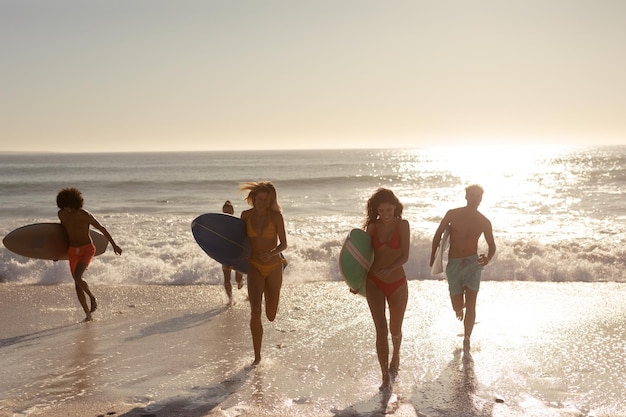 Многоэтническая группа друзей мужского и женского пола на отдыхе на пляже с досками для серфинга, выбегающими из моря на закате