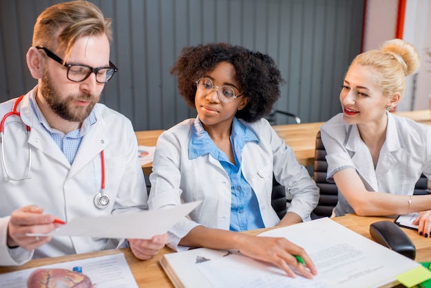 Foto gruppo multietnico di studenti di medicina in uniforme che discutono seduti insieme alla scrivania con diverse cose mediche in classe