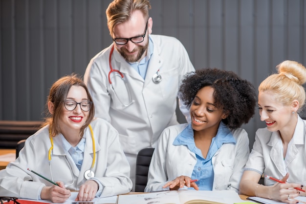 Gruppo multietnico di studenti di medicina in uniforme che studiano insieme seduti alla scrivania con libri nell'aula moderna