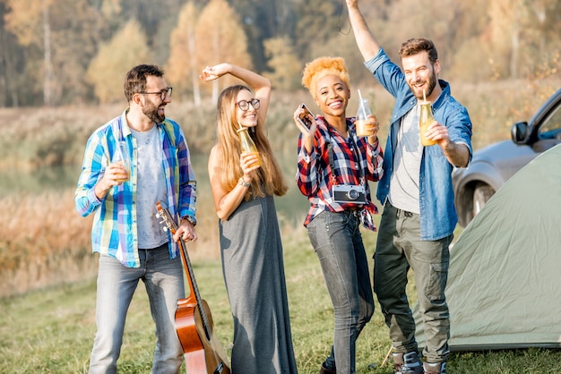 Фото Многонациональная группа друзей, одетых в повседневную одежду, весело танцует с напитками во время отдыха на природе в кемпинге возле леса