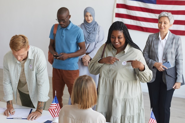 Gruppo multietnico di persone che si registrano al seggio elettorale decorato con bandiere americane il giorno delle elezioni, concentrarsi sulla donna africana sorridente che punta all'adesivo i voto, spazio di copia