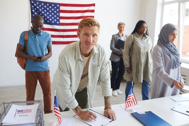 Gruppo multietnico di persone che si registrano al seggio elettorale decorato con bandiere americane il giorno delle elezioni, concentrarsi sul giovane che firma moduli di voto e copia spazio