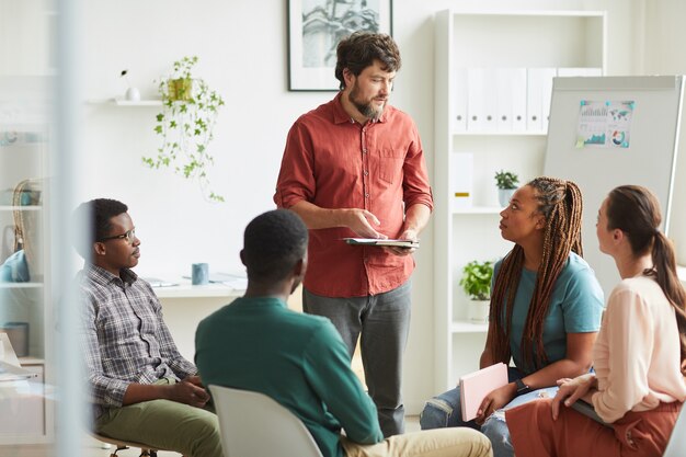 Multi-ethnic group of people sitting in circle while discussing business project in office, focus on smiling beaqrded manager talking to colleagues