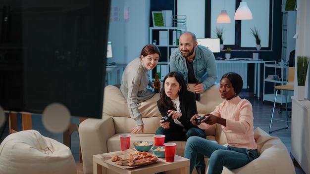 Multi ethnic group of people using joysticks to play video\
games on tv console for entertainment after work. workmates playing\
game with controllers on television to do fun activity.