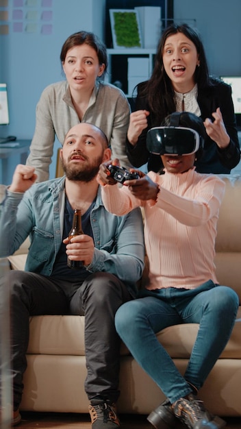 Multi ethnic people cheering while woman winning game with vr\
glasses and controller on tv console. workmates enjoying video\
games with joystick to have fun with activity after work