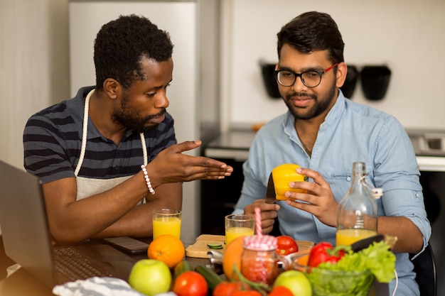 Studenti multietnici seduti a tavola in cucina a guardare la ricetta video sul laptop e imparano a cucinare cibi sani. ragazzo africano in grembiule con il suo amico indiano che prepara insalata, affettando il peperone.