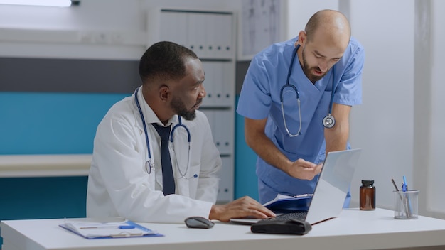 Multi-ethnic team discussing healthcare treatment analyzing sickness expertise documents working in hospital office. African american therapist doctor typing medical report on laptop computer