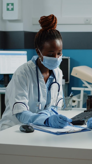 Multi ethnic team of doctor and nurse preparing checkup document for examination with black patient on bed. Diverse medical specialists filling in prescription paper for ill person