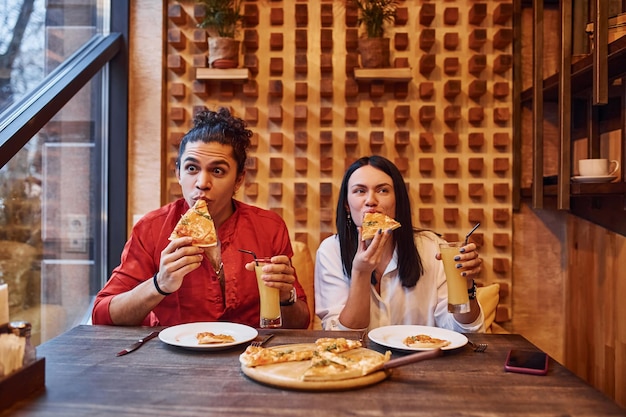 Multi ethnic young couple sitting indoors together and eating pizza.