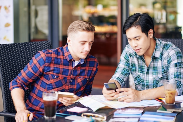 Multi-ethnic young people in plaid shirts meeting at cafe table and discussing new travel app on smartphone