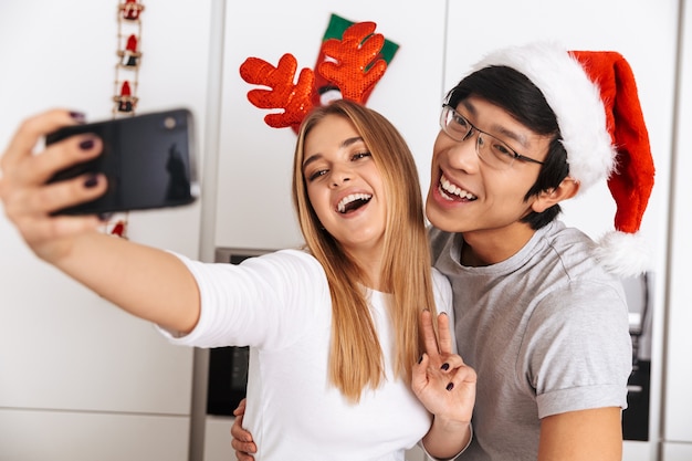 multi-etnisch stel, man en vrouw die kerst outfit dragen, permanent in lichte keuken en selfie foto nemen op mobiele telefoon
