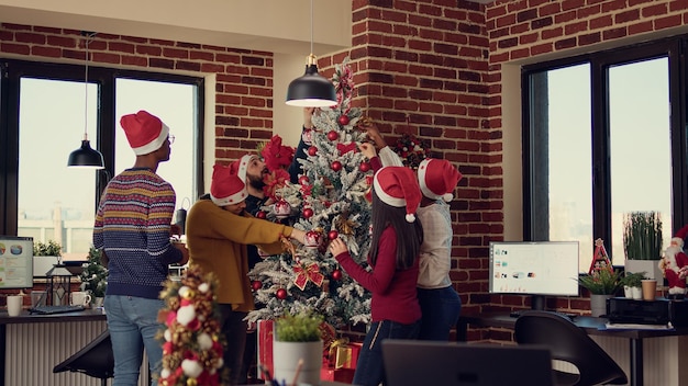 Multi-etnisch team van mensen die een kerstboomdecor in een feestelijk kantoor zetten, voorbereiden op de winterseizoentraditie op het werk. Werkplek versieren met kerstversieringen voor seizoensgebonden vakantie.