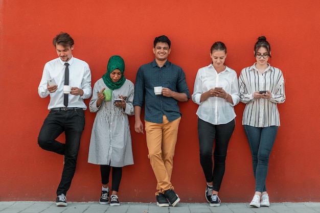 Multi-etnische groep informele zakenmensen die smartphones gebruiken tijdens een koffiepauze van het werk voor de rode muur buiten. Foto van hoge kwaliteit