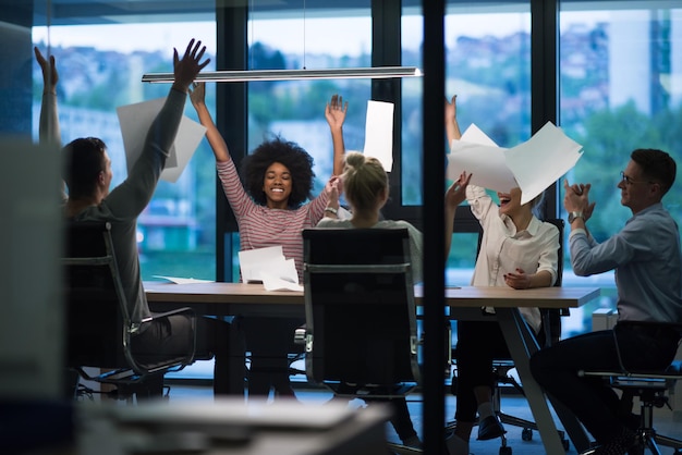 multi-etnische groep jonge zakenmensen die documenten gooien en er gelukkig uitzien terwijl ze succes vieren op hun werkplek in het nachtkantoor