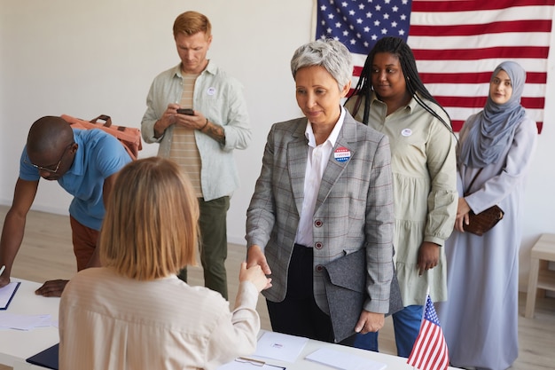 Multi-etnische groep mensen bij het stembureau versierd met Amerikaanse vlaggen op verkiezingsdag, focus op lachende senior vrouw handen schudden met stemmend ambtenaar, kopie ruimte