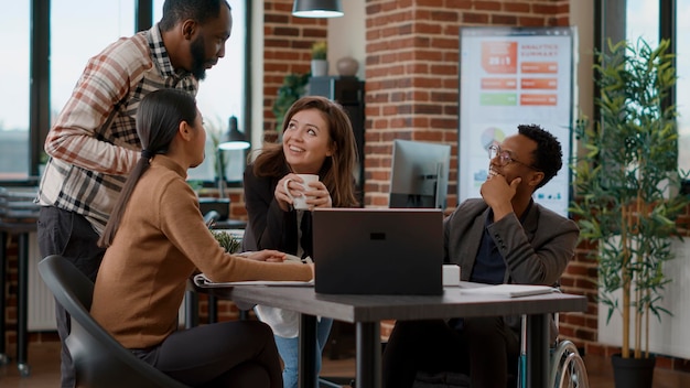 Multi-etnische groep mensen die lachen en plezier hebben op het werk, ontmoeten elkaar om over de opdracht te praten. Collega's die zich gelukkig voelen en samenwerken om de groei in een gehandicaptenvriendelijk kantoor te analyseren.