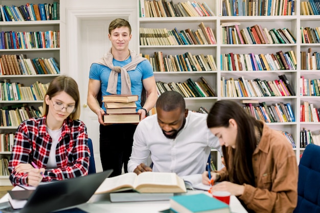 Multi-etnische groep van jonge mensen, studenten, samen studeren aan tafel, boeken lezen. De jonge stapel van de jongensholding vele boeken bevindt zich achter de lijst en bekijkt zijn vrienden