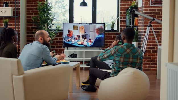 Multi-etnische groep vrienden die films kijken op tv-kanaal, bier drinken en snacks eten tijdens een leuke bijeenkomst. Gelukkige mensen die thuis genieten van tv-show of bioscoopfilm op televisieprogramma.