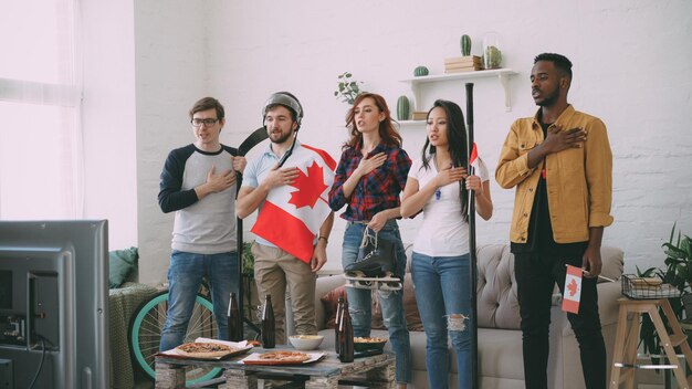 Multi-etnische groep vrienden die het Canadese volkslied luisteren en zingen voordat ze naar sport kijken