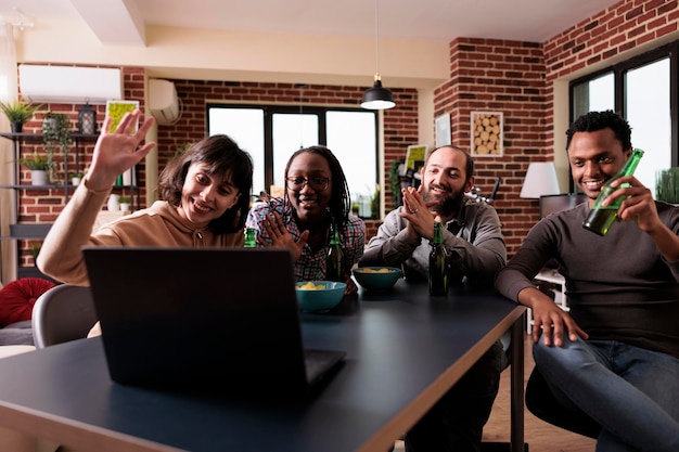 Multi-etnische groep vrienden die met de hand zwaaien tijdens online videogesprekconferentie op laptopcomputer. Diverse mensen zitten thuis in de woonkamer terwijl ze een begroetingsgebaar maken tijdens een internetgesprek.