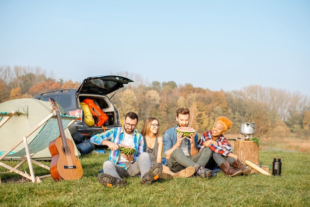 Multi-etnische groep vrienden die picknicken, watermeloen eten, op een rij zitten op de camping met tent, auto en wandeluitrusting in de buurt van het meer