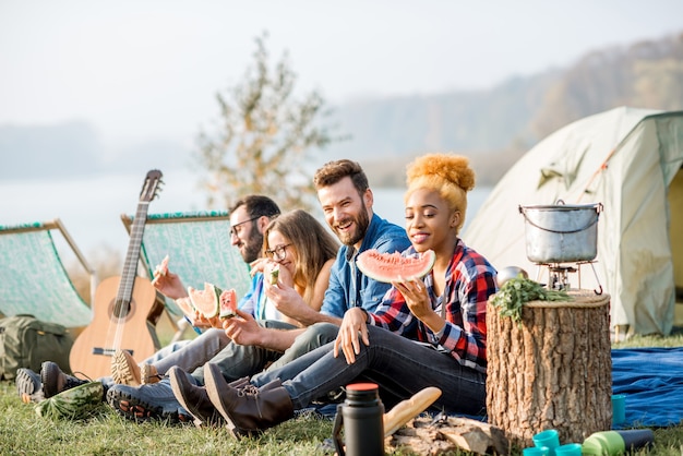 Multi-etnische groep vrienden die picknicken, watermeloen eten tijdens de openluchtrecreatie met tent, auto en wandeluitrusting in de buurt van het meer