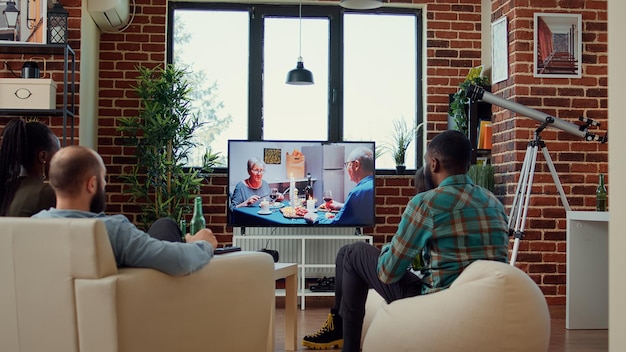 Multi-etnische groep vrienden genieten van film op tv-kanaal, bier drinken en snacks eten. Gelukkige mensen kijken naar dramafilm of tv-show op televisieprogramma tijdens een leuke thuisbijeenkomst.