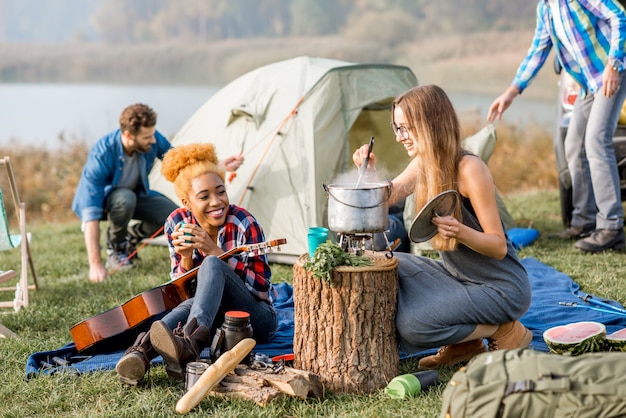 Multi-etnische groep vrienden terloops gekleed met een picknick, soep koken met ketel tijdens de openluchtrecreatie in de buurt van het meer