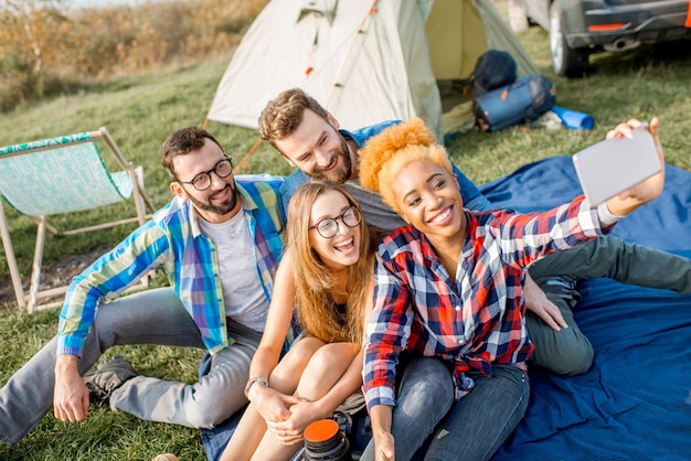 Multi-etnische groep vrienden terloops gekleed met plezier samen een selfie-foto maken tijdens de openluchtrecreatie met tent, auto en wandeluitrusting in de buurt van het meer