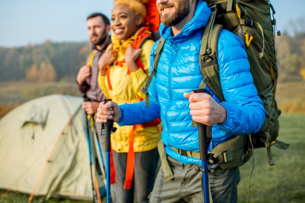 Multi-etnische vrienden in kleurrijke jassen wandelen met rugzakken, staande in de buurt van de camping op het groene gazon