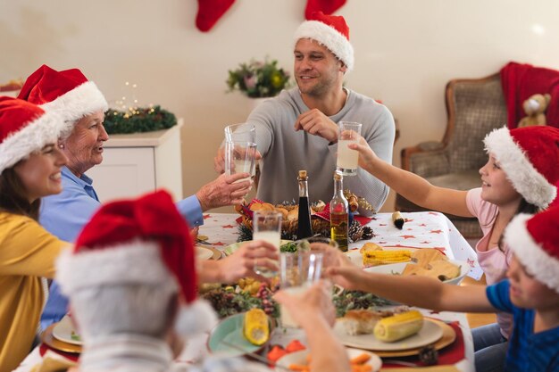 Famiglia caucasica di più generazioni seduta a tavola per cenare insieme, indossando cappelli da babbo natale, facendo un brindisi e sorridendo. celebrazione natalizia di qualità in famiglia.