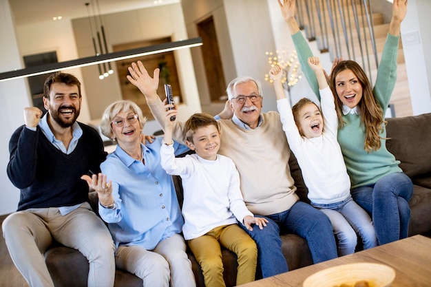 Famiglia multigenerazionale che guarda un calcio in tv e celebra un goal, seduta sul divano in soggiorno