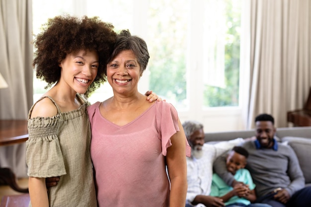 Multi-generation mixed-race family enjoying their time at home together, sitting on a couch, senior woman and her daughter are standing, embracing, looking at the camera and smiling with their  family