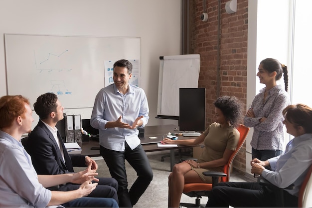 Multi racial businesspeople gather at briefing positive team leader talking to office employees interns or students at corporate training teach explain project strategy discuss business plan concept