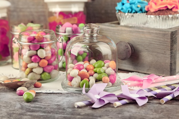 Multicolor candies in glass jars and cupcakes on wooden background