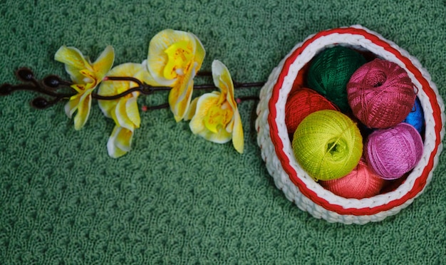 multicolored balls of thread in wicker basket on green crocheted background Yellow branch of orchid