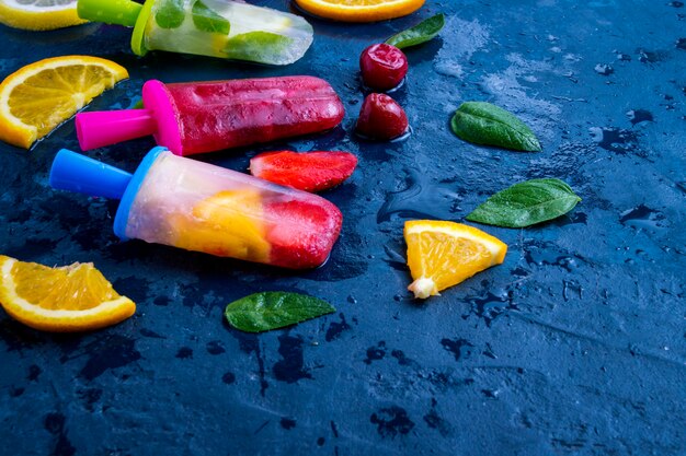 Multicolored bright fruit popsicle with strawberry, cherry, lemon, orange, lemon and mint aroma and fresh fruit on a dark blue surface