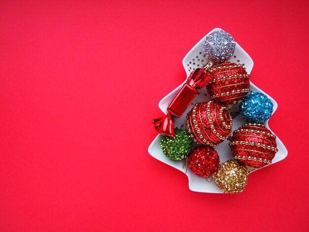 Multicolored Christmas balls on a white plate in the form of a Christmas tree Christmas tree on the right Copy space on the left Red background Beautiful striped Christmas decor and sparkles