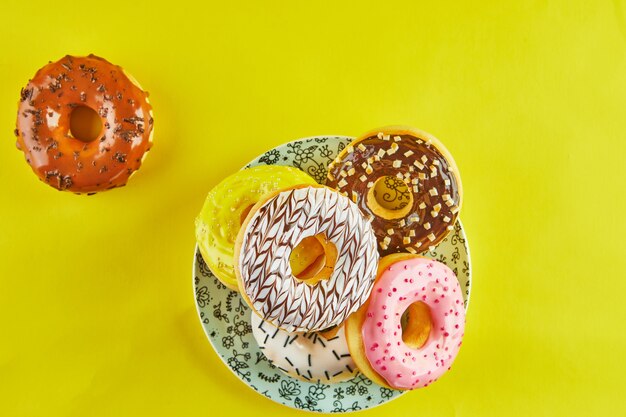 Multicolored donuts with icing and splashes on a blue plate on a yellow background. flat lay