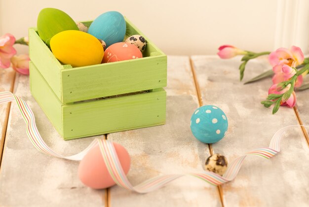 Multicolored Easter eggs in a wooden box, ribbons and flowers on a wooden.