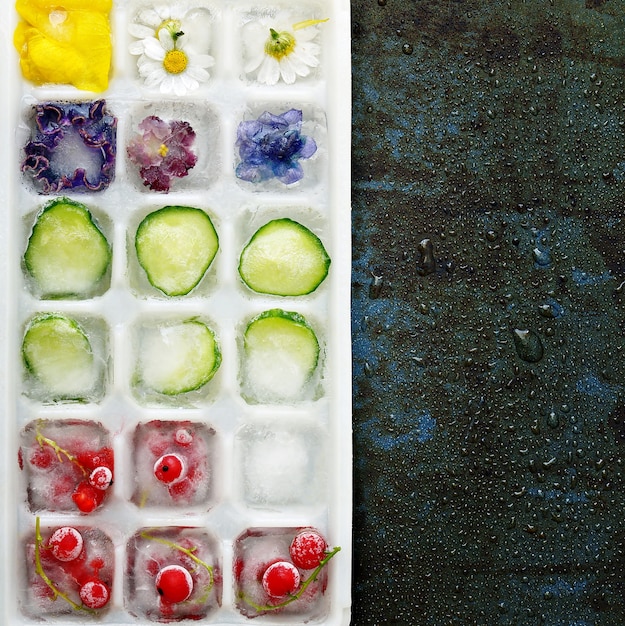 Cubetti di ghiaccio congelati multicolori con frutta, fiori e verdure su uno sfondo scuro sotto forma di ghiaccio