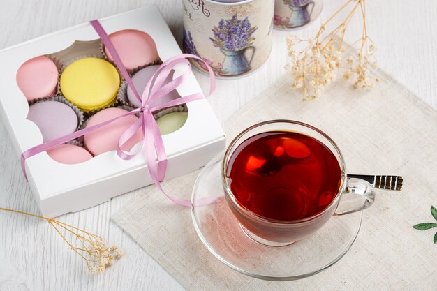 Multicolored macaroons in a box and a cup of tea on a light wood kitchen table