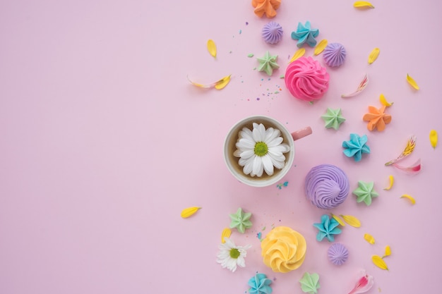 Multicolored marshmallows with a mug of tea and flowers on a pink background. Summer mood. View from above. space for text.