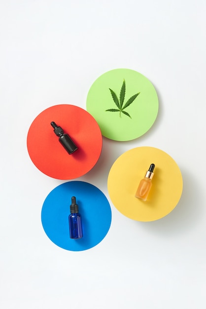 ライトグレーのテーブル、コピースペースに天然医療大麻エッセンシャルCBDオイルのボトルが入った色とりどりの紙の丸いカード。医療目的での大麻の使用。上面図。