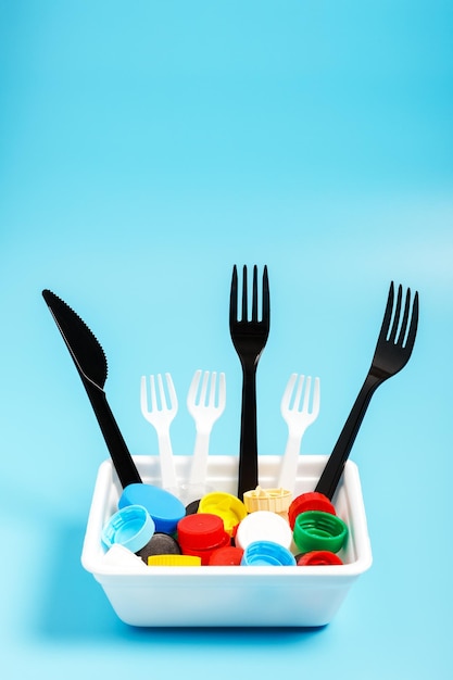 Multicolored plastic bottle caps and plastic forks in a white plastic box on a blue background The problem of environmental pollution Recycling plastic