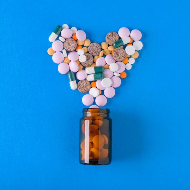 Multicolored tablets are poured from a glass vessel in the shape of a heart on a blue background. The view from the top. The concept of treatment and prevention of diseases. Flat lay.